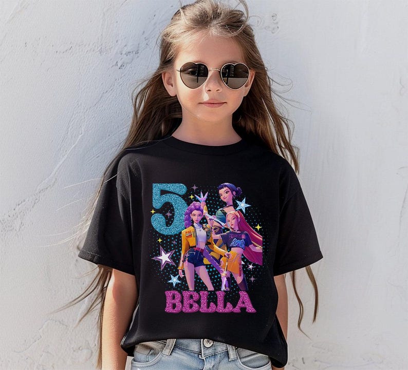 May include: Black t-shirt featuring a colorful graphic design with the number "5" in blue glitter and the name "BELLA" in pink glitter. The graphic includes illustrations of animated characters. The shirt is worn by a young person.