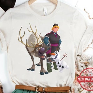 May include: White t-shirt featuring a cartoon illustration of a reindeer, a snowman, and a man with brown hair wearing a black and purple outfit. The man is holding a staff and the reindeer is looking to the right. The snowman is smiling and has two black eyes.