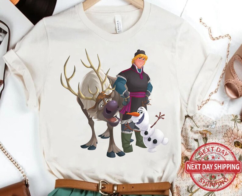 May include: White t-shirt featuring a cartoon illustration of a reindeer, a snowman, and a man with brown hair wearing a black and purple outfit. The man is holding a staff and the reindeer is looking to the right. The snowman is smiling and has two black eyes.
