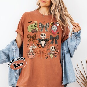 May include: A burnt orange t-shirt with a design featuring characters and bows from The Nightmare Before Christmas. The design includes Jack Skellington, Sally, Oogie Boogie, and other characters. The shirt has the words "Comfort Colors" and "Yam" on it.