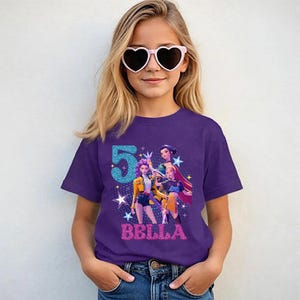May include: A purple t-shirt featuring a graphic of animated characters, the number "5" in blue glitter, and the name "BELLA" in pink glitter. The shirt is worn by a person wearing heart-shaped sunglasses.