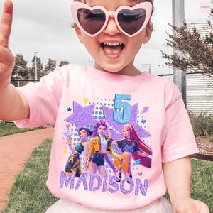 May include: A pink t-shirt featuring a graphic of three animated figures, a large number 5, and the name "MADISON" in purple glitter. The shirt is worn by a child wearing heart-shaped sunglasses.