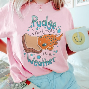 May include: A pink t-shirt featuring a cartoon fish with a peanut butter and jelly sandwich on its head. The text on the shirt reads "Pudge controls the weather".