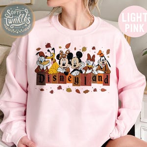 May include: A light pink sweatshirt with a vintage-style graphic of Mickey Mouse, Minnie Mouse, Donald Duck, Goofy, and Pluto. The characters are standing in front of a sign that says "Disneyland" with autumn leaves scattered around them.