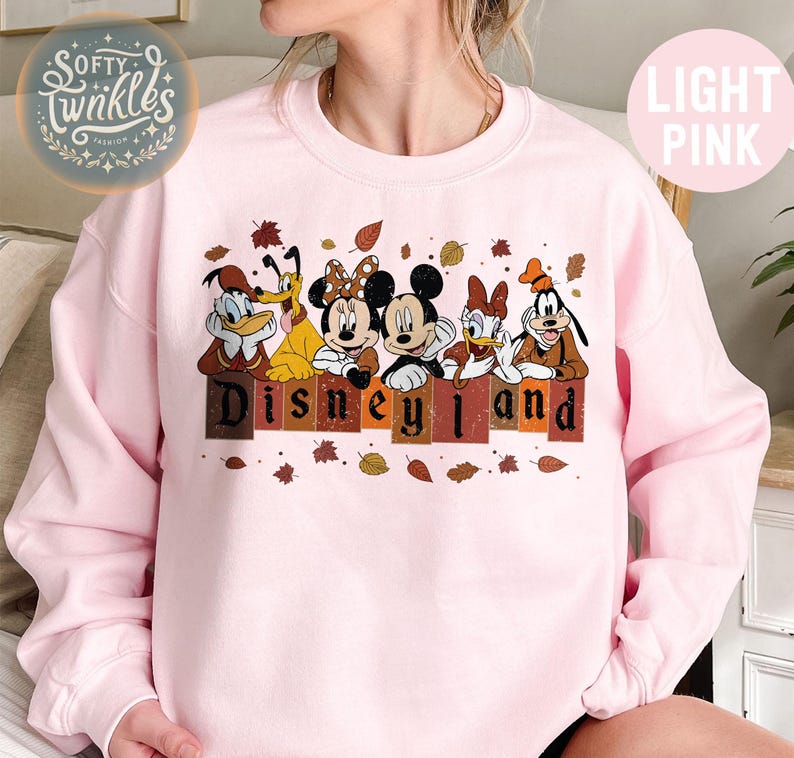 May include: A light pink sweatshirt with a vintage-style graphic of Mickey Mouse, Minnie Mouse, Donald Duck, Goofy, and Pluto. The characters are standing in front of a sign that says "Disneyland" with autumn leaves scattered around them.
