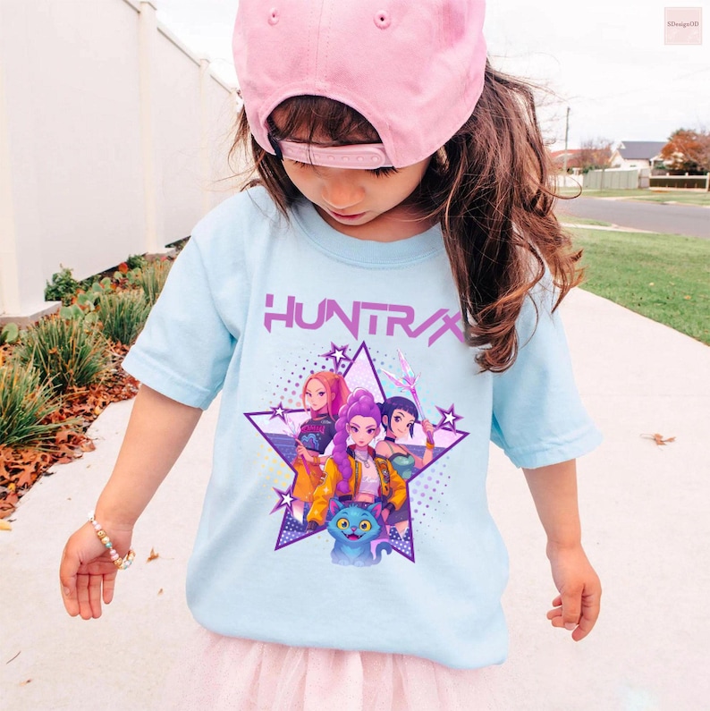 Pode incluir: Camiseta azul clara com a palavra "HUNTRA" em roxo, e um gr&aacute;fico de tr&ecirc;s mulheres animadas e um gato azul dentro de uma estrela roxa. A camiseta &eacute; usada por uma crian&ccedil;a usando um bon&eacute; de beisebol rosa.