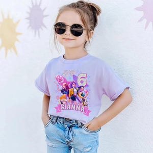 May include: A light purple t-shirt featuring a colorful graphic of three cartoon characters, musical notes, and the number 6. The name "GIANNA" is printed on a pink banner. The girl is wearing sunglasses and blue jeans.