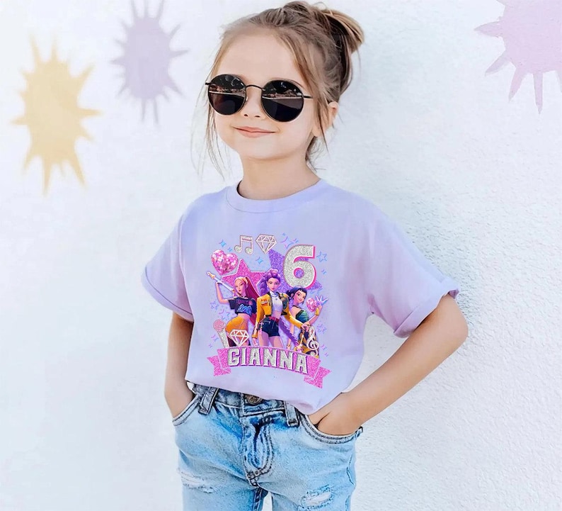 May include: A light purple t-shirt featuring a colorful graphic of three cartoon characters, musical notes, and the number 6. The name "GIANNA" is printed on a pink banner. The girl is wearing sunglasses and blue jeans.