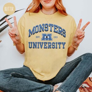 May include: Yellow t-shirt with the text "MONSTERS UNIVERSITY" in blue. The shirt features a collegiate-style design with the words "EST. 1313" and a stylized eye graphic. The shirt is a casual wear item.