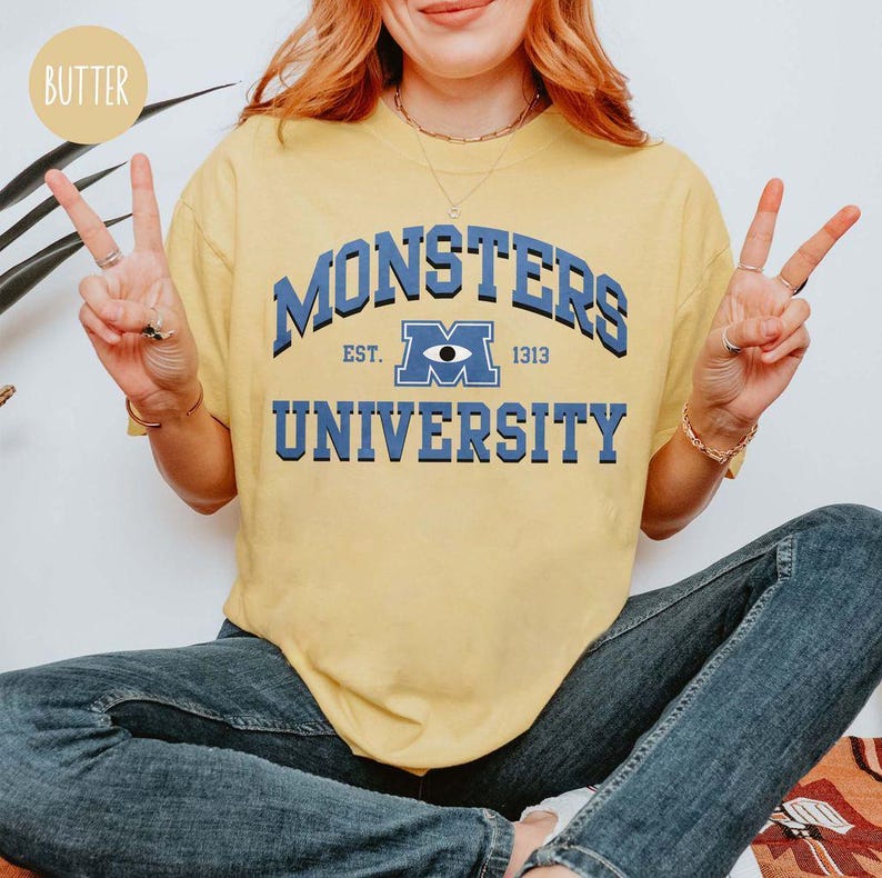 May include: Yellow t-shirt with the text "MONSTERS UNIVERSITY" in blue. The shirt features a collegiate-style design with the words "EST. 1313" and a stylized eye graphic. The shirt is a casual wear item.