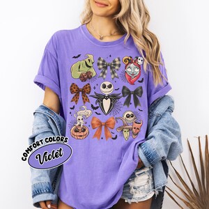 May include: A violet Comfort Colors t-shirt with Halloween character illustrations. The design includes Jack Skellington, Sally, and Oogie Boogie, along with bats and bows. The shirt has short sleeves and the text "Comfort Colors Violet".