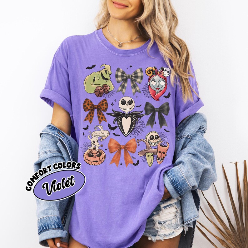 May include: A violet Comfort Colors t-shirt with Halloween character illustrations. The design includes Jack Skellington, Sally, and Oogie Boogie, along with bats and bows. The shirt has short sleeves and the text "Comfort Colors Violet".