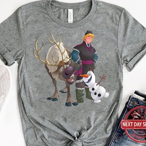 May include: A grey t-shirt featuring a cartoon image of a reindeer, Olaf the snowman, and a man with brown hair and a blue shirt. The man is wearing a purple sash and holding a staff. The reindeer is brown and white and has antlers. Olaf is a snowman with a carrot nose and a stick body. The t-shirt has a grey background.