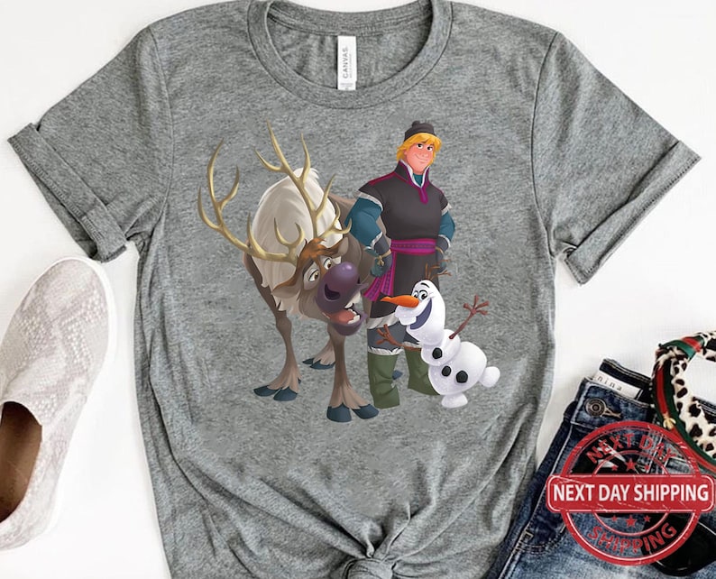 May include: A grey t-shirt featuring a cartoon image of a reindeer, Olaf the snowman, and a man with brown hair and a blue shirt. The man is wearing a purple sash and holding a staff. The reindeer is brown and white and has antlers. Olaf is a snowman with a carrot nose and a stick body. The t-shirt has a grey background.