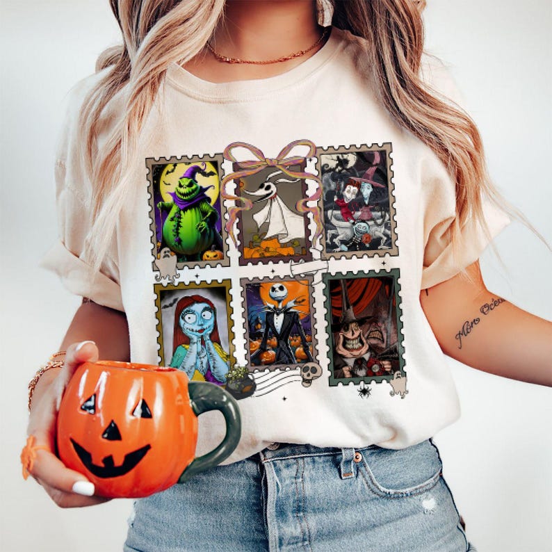 May include: Cream-colored t-shirt featuring six postage stamp-style images of characters from The Nightmare Before Christmas. The shirt is paired with an orange pumpkin-shaped mug. The mug has a black handle and a carved jack-o'-lantern face.