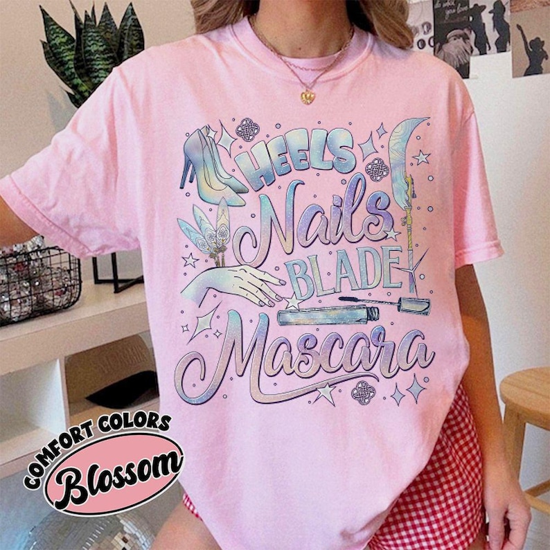 May include: A light pink t-shirt with the words "Heels, Nails, Blade, Mascara" in a cursive font. The shirt features illustrations of a high heel, a hand with a nail, a blade, and a mascara tube. The words "Comfort Colors" and "Blossom" are also printed on the shirt.