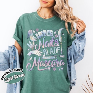 May include: Light green Comfort Colors t-shirt with a graphic design featuring the words "Heels, Nails, Blade, Mascara" in a pastel color scheme. The design includes illustrations of a high heel, hand, and makeup items.