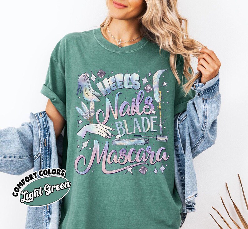 May include: Light green Comfort Colors t-shirt with a graphic design featuring the words "Heels, Nails, Blade, Mascara" in a pastel color scheme. The design includes illustrations of a high heel, hand, and makeup items.