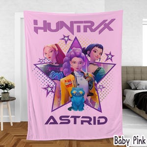 Könnte beinhalten: Eine babyrosa Decke mit einer Grafik von drei animierten Frauen in einem Stern, mit dem Text "HUNTRAX" oben und "ASTRID" unten. In der Mitte befindet sich eine blaue Katze. Die Decke wird in einem Schlafzimmer gezeigt.