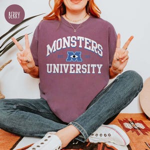 May include: Berry-colored sweatshirt with the text "MONSTERS UNIVERSITY" in white and blue. The sweatshirt features a graphic of the Monsters University logo. The person is wearing blue jeans and white sneakers.