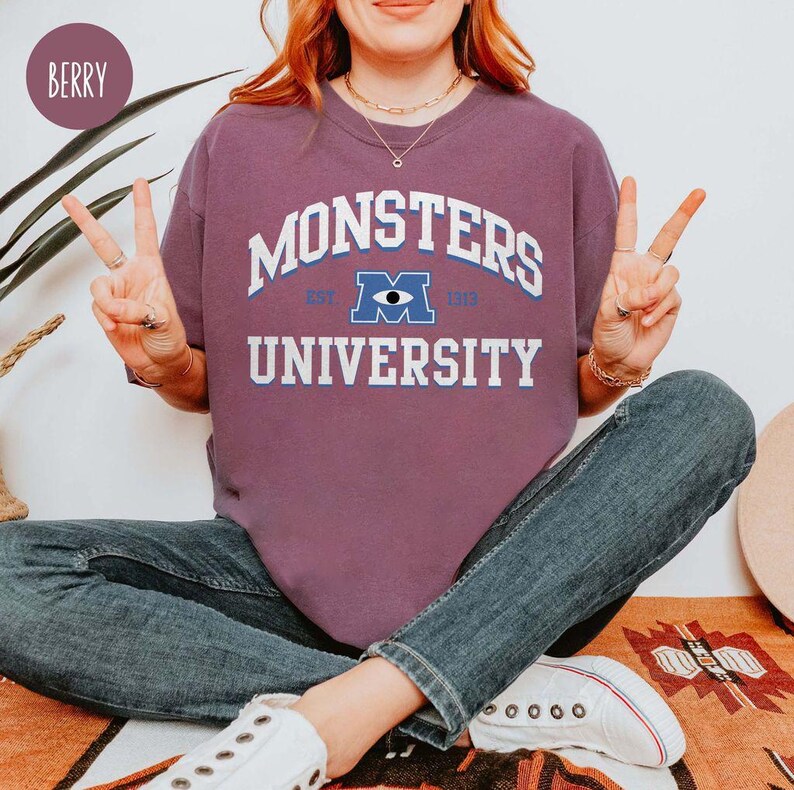 May include: Berry-colored sweatshirt with the text "MONSTERS UNIVERSITY" in white and blue. The sweatshirt features a graphic of the Monsters University logo. The person is wearing blue jeans and white sneakers.