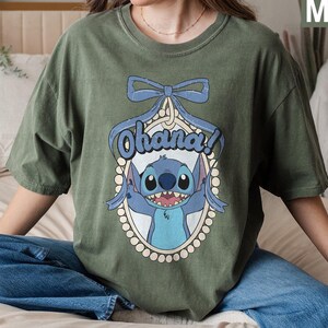 Puede incluir: Una camiseta verde con un personaje azul de Stitch de Lilo & Stitch. El personaje sostiene un cartel que dice "Ohana!" y est&aacute; rodeado por un borde de perlas. La camiseta tiene un dise&ntilde;o de lazo en la parte superior.