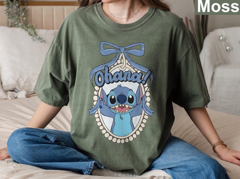 Puede incluir: Una camiseta verde con un personaje azul de Stitch de Lilo & Stitch. El personaje sostiene un cartel que dice "Ohana!" y est&aacute; rodeado por un borde de perlas. La camiseta tiene un dise&ntilde;o de lazo en la parte superior.