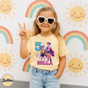 May include: A light yellow t-shirt with a graphic of three animated characters, the number 5, and the name "BELLA" in pink glitter. The girl is wearing heart-shaped sunglasses and blue jeans. The background features rainbows and smiling suns.