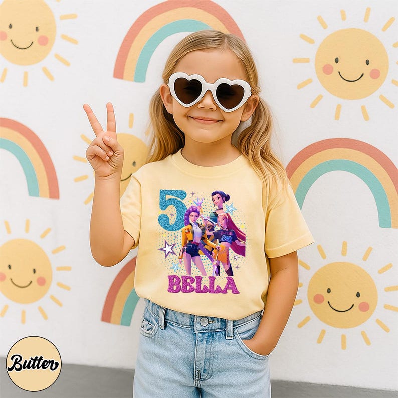 May include: A light yellow t-shirt with a graphic of three animated characters, the number 5, and the name "BELLA" in pink glitter. The girl is wearing heart-shaped sunglasses and blue jeans. The background features rainbows and smiling suns.