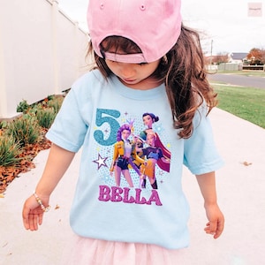 May include: A light blue t-shirt featuring a graphic of three animated characters, the number "5" in teal glitter, and the name "BELLA" in pink glitter. The shirt is worn by a child wearing a pink baseball cap.