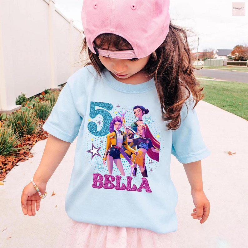 May include: A light blue t-shirt featuring a graphic of three animated characters, the number "5" in teal glitter, and the name "BELLA" in pink glitter. The shirt is worn by a child wearing a pink baseball cap.