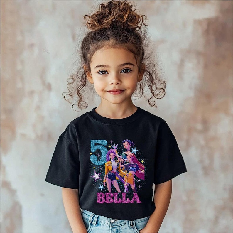 May include: A black t-shirt featuring a colorful graphic of three animated characters and the number 5 in blue glitter. The name "BELLA" is written in pink glitter below the graphic. The shirt is worn by a young girl.