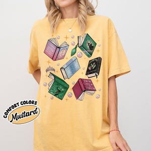 May include: Mustard-colored t-shirt featuring open books with various titles and colors, including pink, green, and blue. The design includes a tiara and bubbles. The words "COMFORT COLORS" and "Mustard" are printed on the shirt.