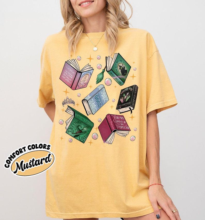 May include: Mustard-colored t-shirt featuring open books with various titles and colors, including pink, green, and blue. The design includes a tiara and bubbles. The words "COMFORT COLORS" and "Mustard" are printed on the shirt.