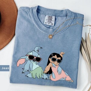 May include: Blue jean t-shirt with a graphic of Stitch and Lilo from the Disney movie Lilo & Stitch. Stitch is wearing sunglasses and a green grass skirt. Lilo is wearing sunglasses and a pink dress. The shirt has a distressed look and is a light blue color.