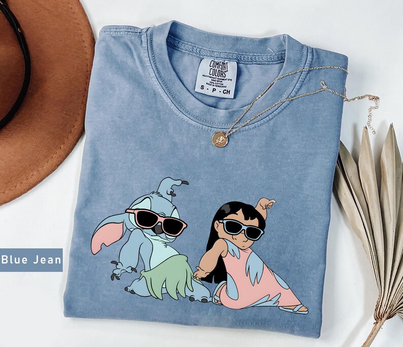 May include: Blue jean t-shirt with a graphic of Stitch and Lilo from the Disney movie Lilo & Stitch. Stitch is wearing sunglasses and a green grass skirt. Lilo is wearing sunglasses and a pink dress. The shirt has a distressed look and is a light blue color.