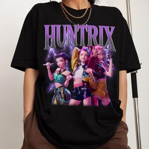 Pu&ograve; includere: T-shirt nera con la scritta "HUNTRIX" in un design viola e argento con un fulmine. La maglietta presenta una grafica di tre donne animate che tengono dei microfoni.