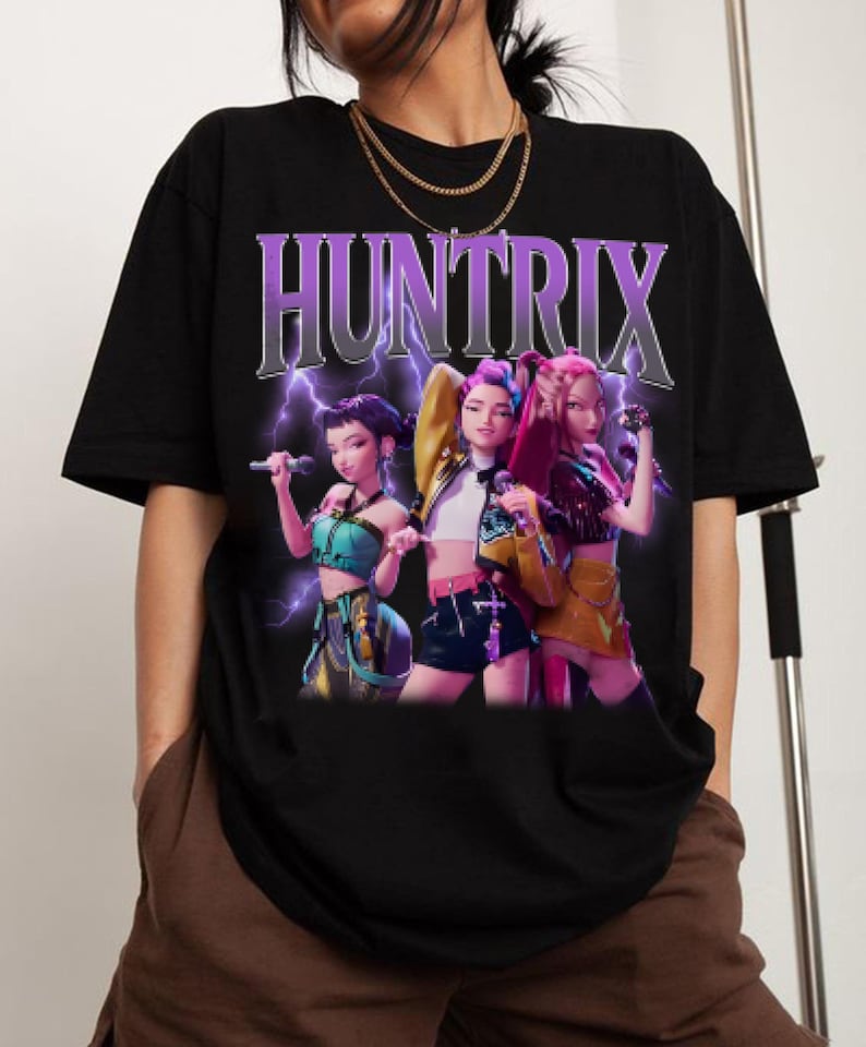 Pu&ograve; includere: T-shirt nera con la scritta "HUNTRIX" in un design viola e argento con un fulmine. La maglietta presenta una grafica di tre donne animate che tengono dei microfoni.