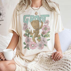 May include: A cream-coloured t-shirt featuring a graphic print of a green character called "Groot" from the Guardians of the Galaxy film franchise. The character is surrounded by pink and green flowers.
