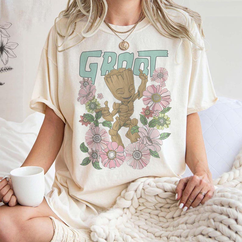 May include: A cream-coloured t-shirt featuring a graphic print of a green character called "Groot" from the Guardians of the Galaxy film franchise. The character is surrounded by pink and green flowers.