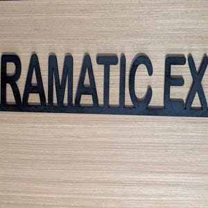 May include: Black metal sign with the words "DRAMATIC EXIT" cut out in a bold, sans-serif font. The sign has a simple, modern design and is mounted on a light wood-grain surface.
