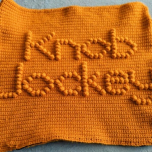 May include: A crocheted throw blanket in a mustard yellow color with the words "Knit Love" in raised lettering.