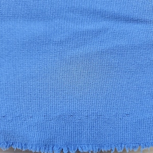 May include: Close-up of a textured, light blue fabric. The fabric has a woven appearance with a slightly frayed edge at the bottom. The color is a solid, bright blue, and the texture appears soft.