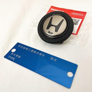 Puede incluir: Un emblema Honda negro y plateado con un símbolo de bocina y una etiqueta TYPE azul. El emblema tiene el logotipo de Honda en el centro. La etiqueta tiene texto japonés y la palabra "TYPE".