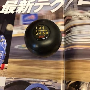 Könnte beinhalten: Ein schwarzer Schalthebelknauf mit einem gelb-roten Gangmuster. Das Gangmuster zeigt die Zahlen 1, 2, 3, 4, 5, 6 und R. Der Schalthebelknauf hat die Buchstaben "STI" in Rot unten.