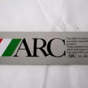 May include: A silver rectangular sticker with the word "ARC" in black letters. The sticker has a green, white, and red stripe design on the left side. The text "The ARC Product is wonders assembled project. They present comfortable unknown car life with ARC power. ARC in JAPAN" is printed on the right side of the sticker.