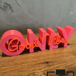 Op de afbeelding: Een roze 3D-geprint bord met de woorden "Good Vibes Only" in oranje script. Het bord staat op een houten oppervlak. De letters zijn dik en blokkerig, met een moderne uitstraling. Het is een decoratief item.