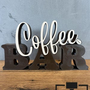 Könnte beinhalten: Ein Holzschild mit der Aufschrift "Coffee Bar". Das Wort "Coffee" ist in weißer Schrift geschrieben, und "Bar" in Blockbuchstaben. Das Schild ist braun mit einem Kaffeebohnen-Detail. Geeignet für die Küche.