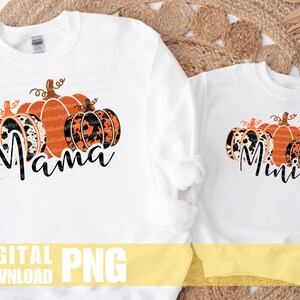 May include: Two white sweatshirts with a pumpkin design. The larger sweatshirt has the word "Mama" and three pumpkins with a black and white animal print. The smaller sweatshirt has the word "Mini" and three pumpkins with a black and white animal print.