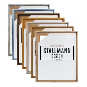 Op de afbeelding: Een verzameling houten fotolijsten in diverse kleuren, waaronder grijs, blauw, bruin en natuurlijke houttinten. De lijsten zijn diagonaal gerangschikt, waarbij hun verschillende afwerkingen worden getoond. De tekst "WOODWORK STALLMANN DESIGN GERMANY" is zichtbaar.